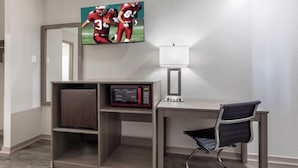 Ein Zimmer mit einem an der Wand befestigten Fernseher, einer Mikrowelle auf einem Regal, einem Schreibtisch mit Stuhl und einer Lampe.
