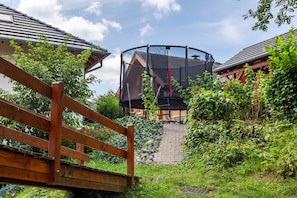 Un pont en bois menant Ă un trampoline dans un espace jardin agrĂ©mentĂ© dâun chemin en briques et de verdure.