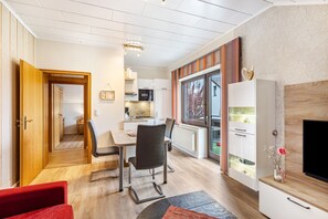 A modern living room with a TV, a dining area with a table and chairs, a kitchen in the background, and a large window. - Haus Schönblick – “Appartment 6” with Mountain View, Balcony and Wi-Fi (Willingen (Upland))