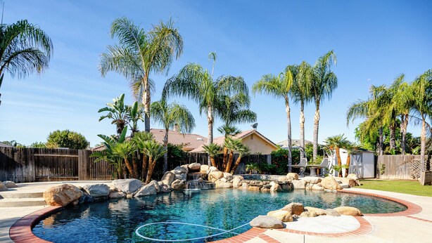 Un patio trasero con piscina, palmeras, una terraza de madera y una casa al fondo.