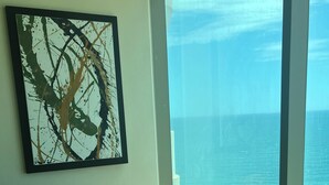 Een slaapkamer met een bed, nachtkastje, een groot abstract schilderij en uitzicht op de oceaan.