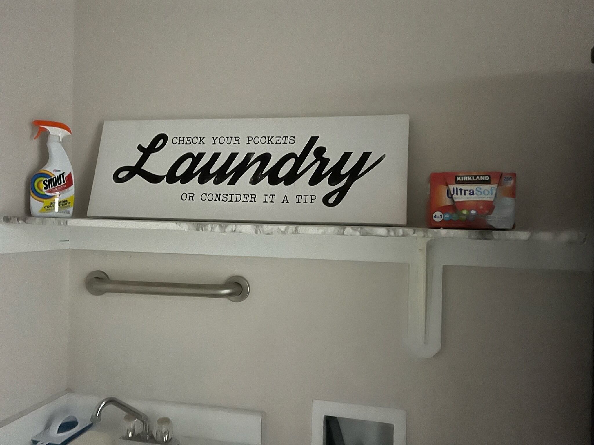 A bathroom with a shelf holding a sign that says "Laundry or consider it a tip" and a bottle of Shout cleaner.