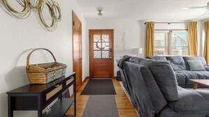 Ein Wohnzimmer mit grauem Sofa, einer Holztür, einem Korbwäschesessel auf einem schwarzen Konsolentisch und Blick durch ein Fenster mit gelben Vorhängen.