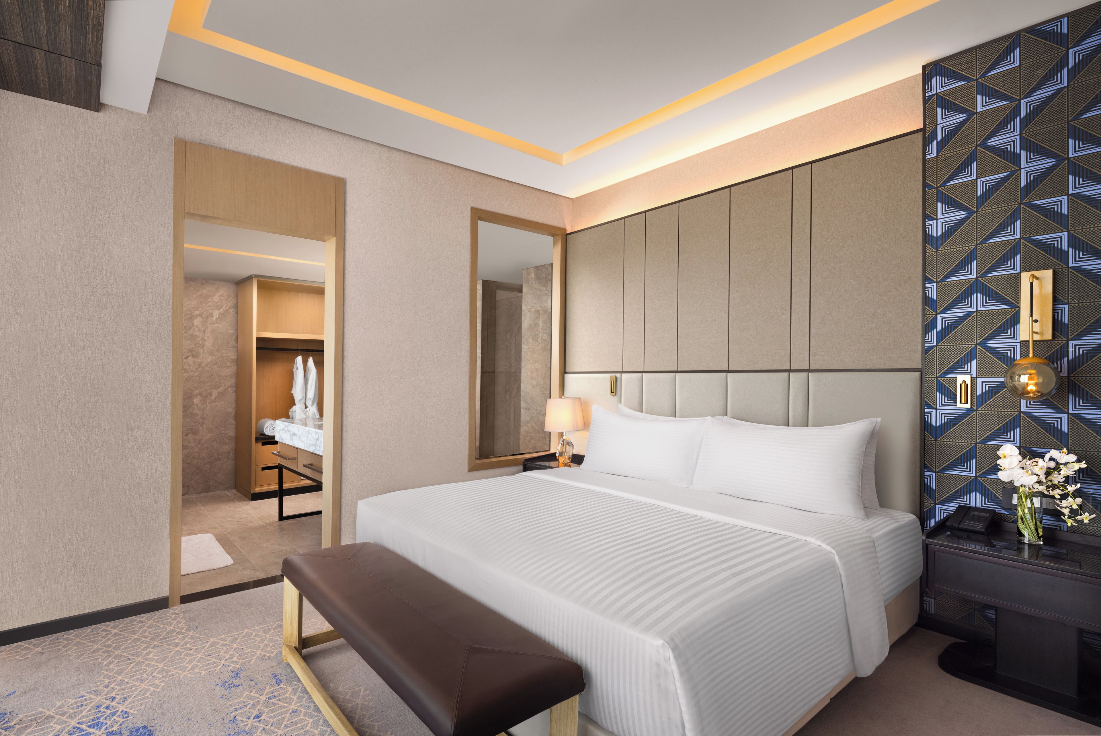 A hotel room with a large bed, a bench, a bedside table with a lamp, and a mirror.