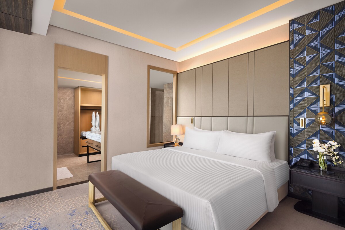 A hotel room with a large bed, a bench, a bedside table with a lamp, and a mirror.