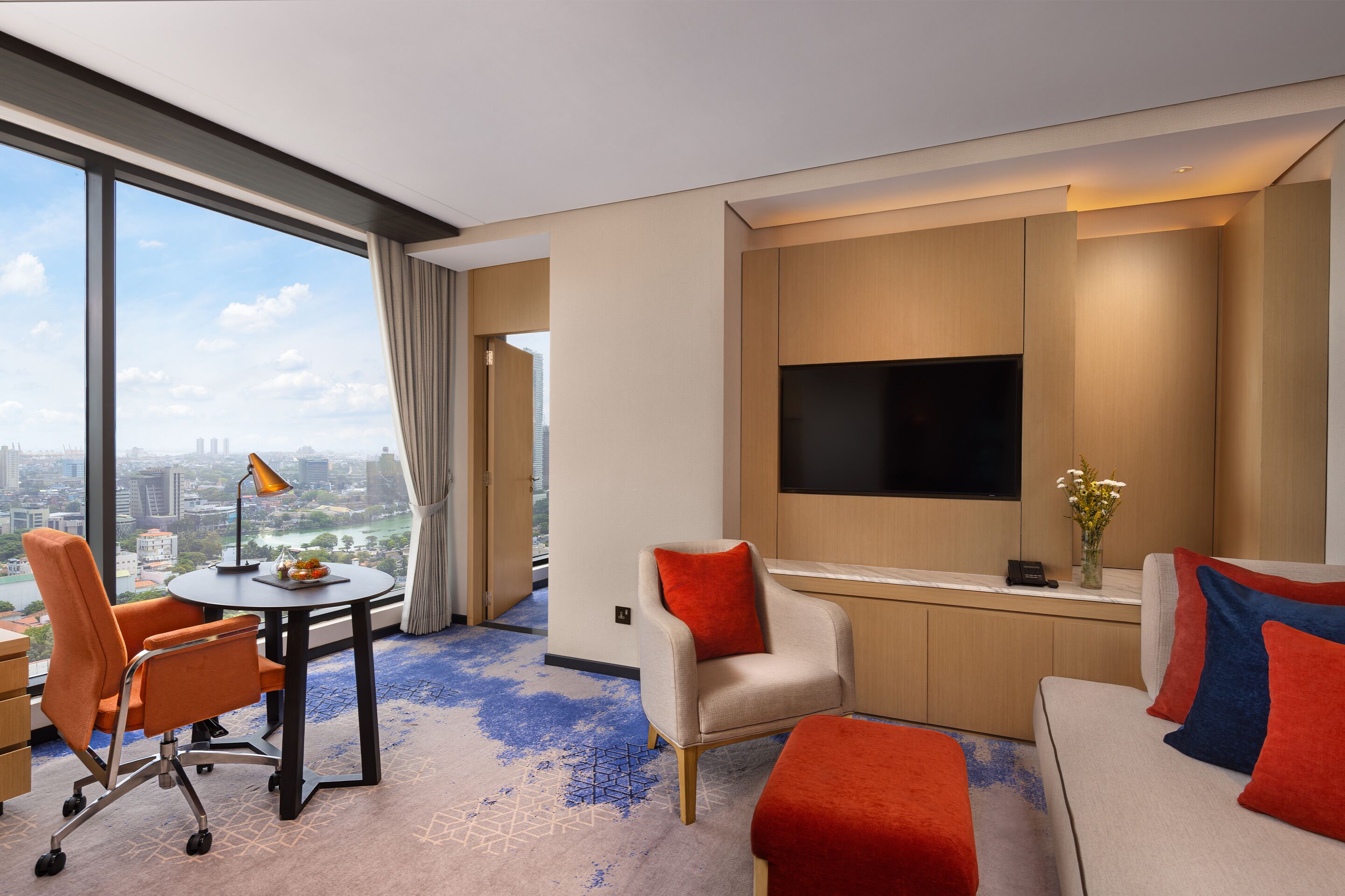 A hotel room with a large window, a flat-screen TV, a sofa, a chair, and a small table.