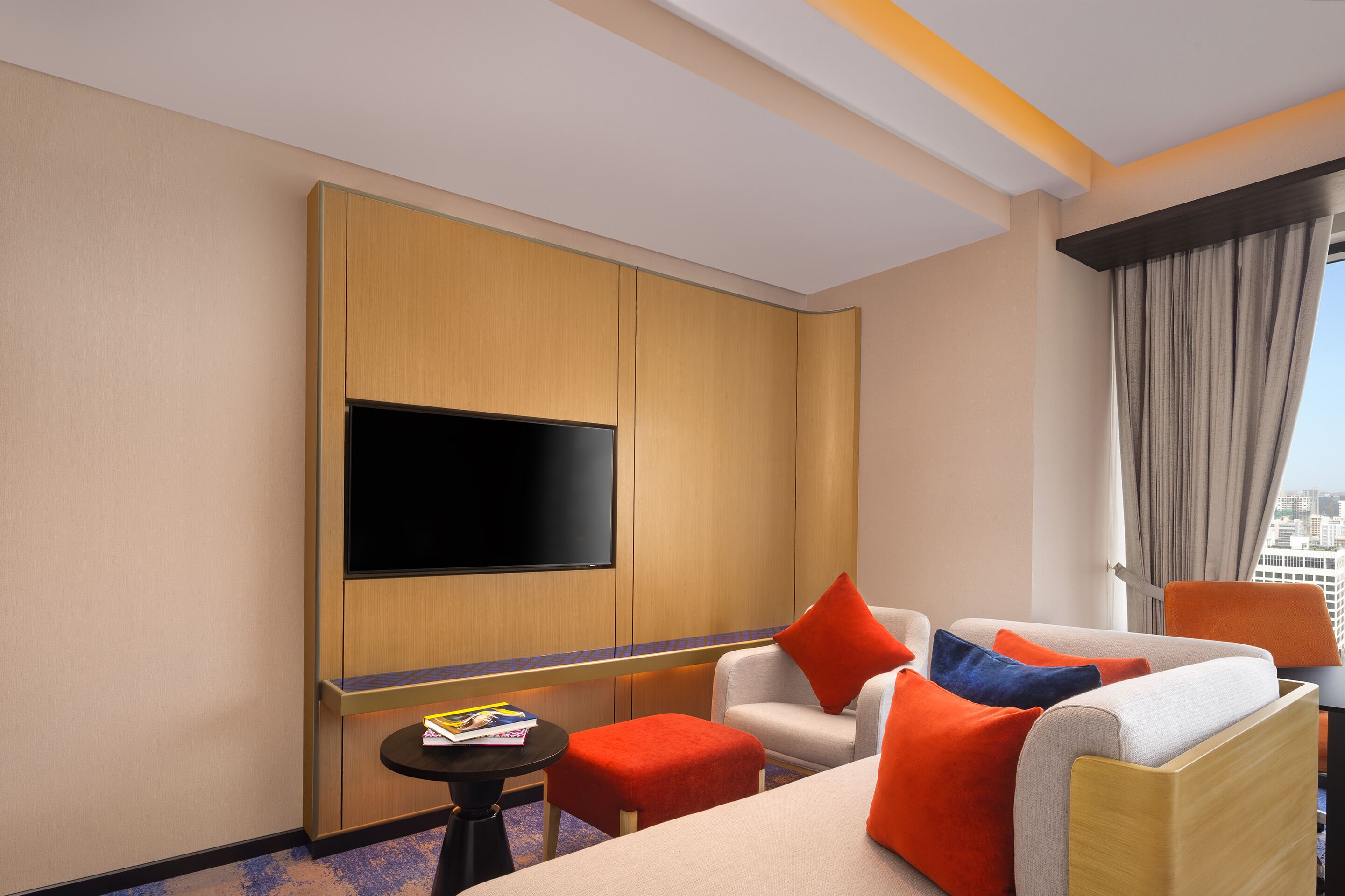 A hotel room with a flat-screen TV, a sofa with orange and blue cushions, a small round table, and a window with curtains.