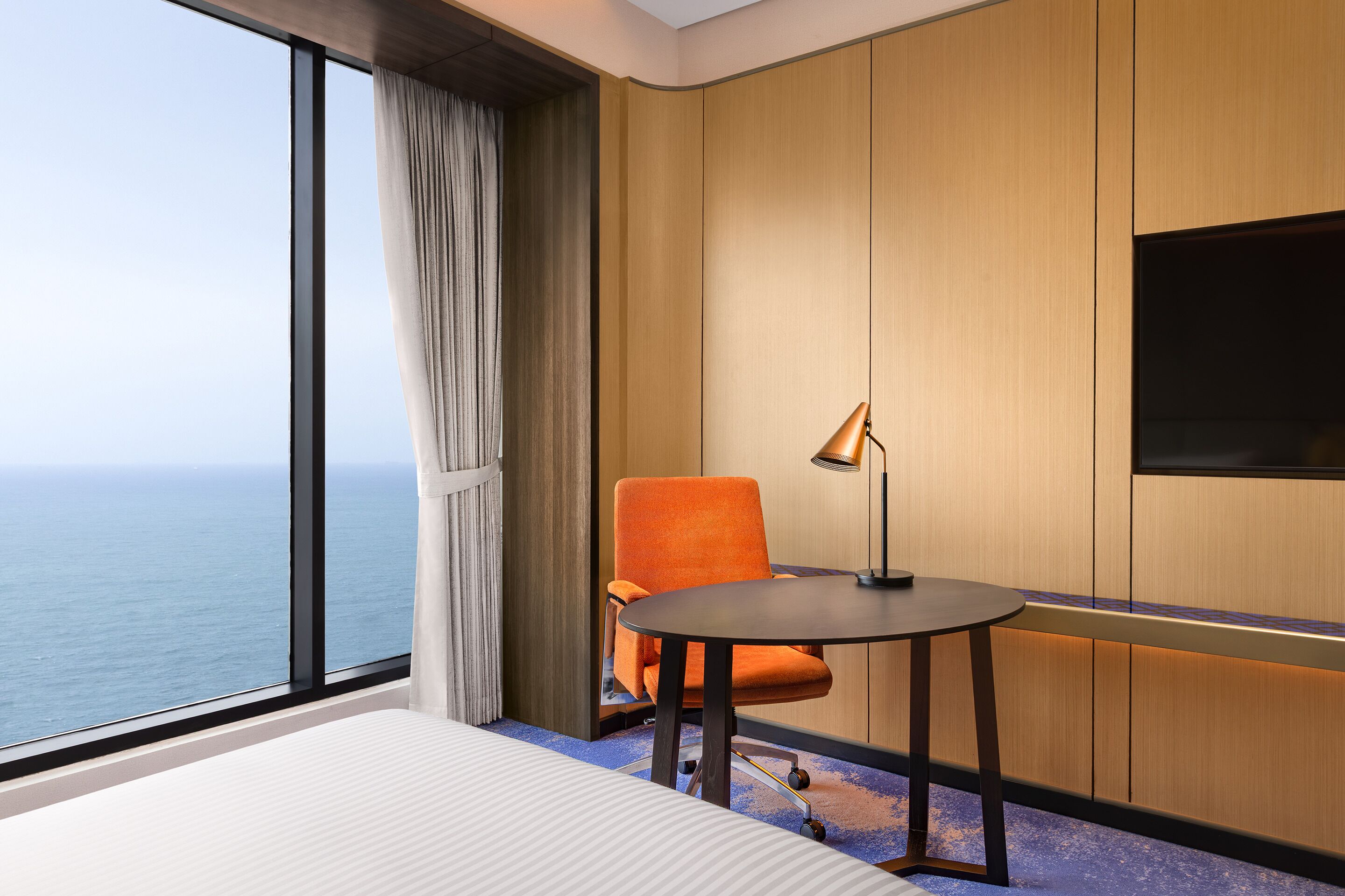 A hotel room with a large window, a bed, an orange chair, a small table, and a TV.