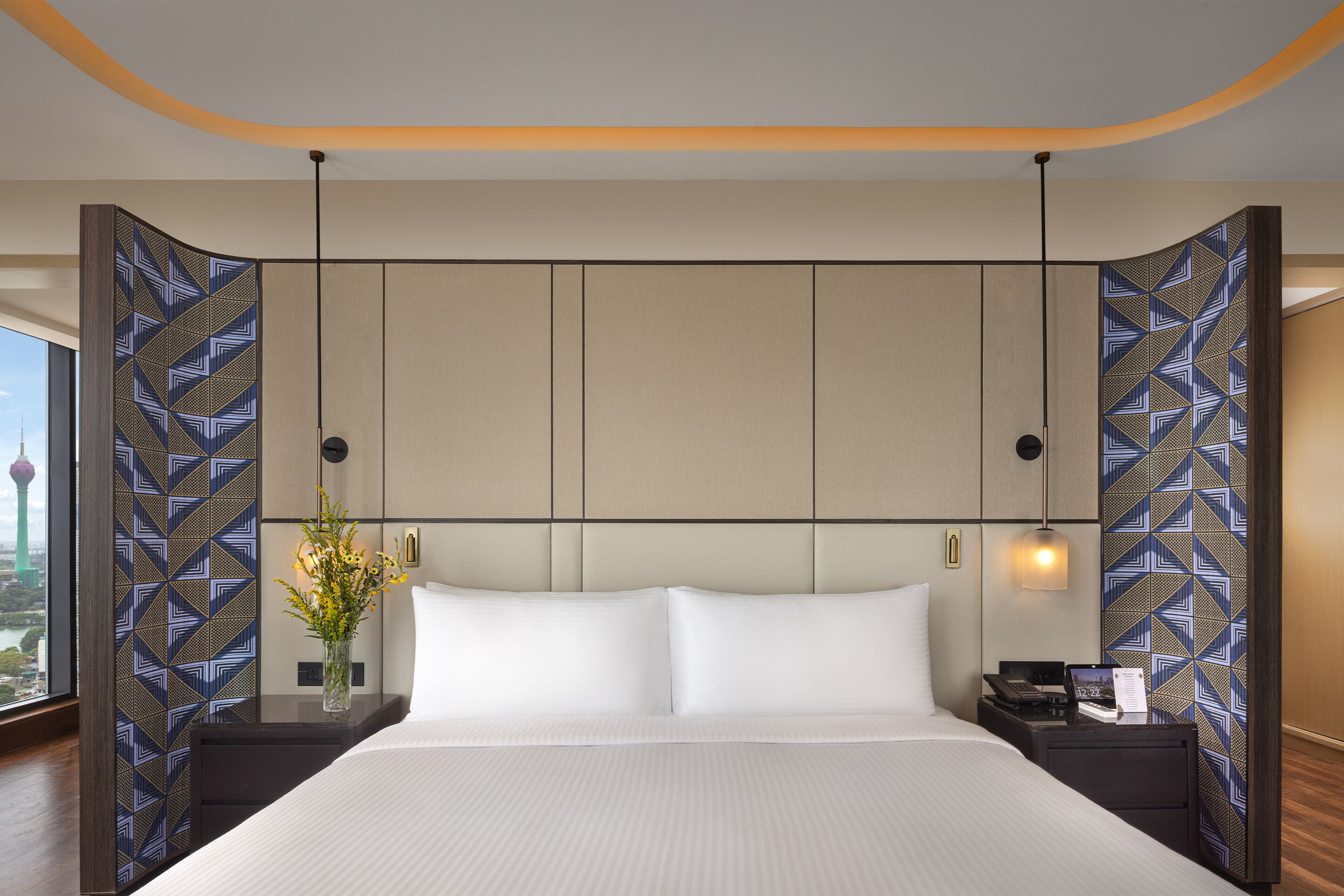 A modern hotel room with a large bed, a nightstand, a vase with flowers, and a city view.