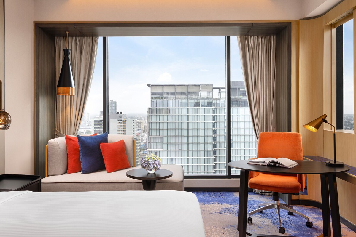 A hotel room with a large window, a sofa, an orange chair, a small table with a book, and a lamp.