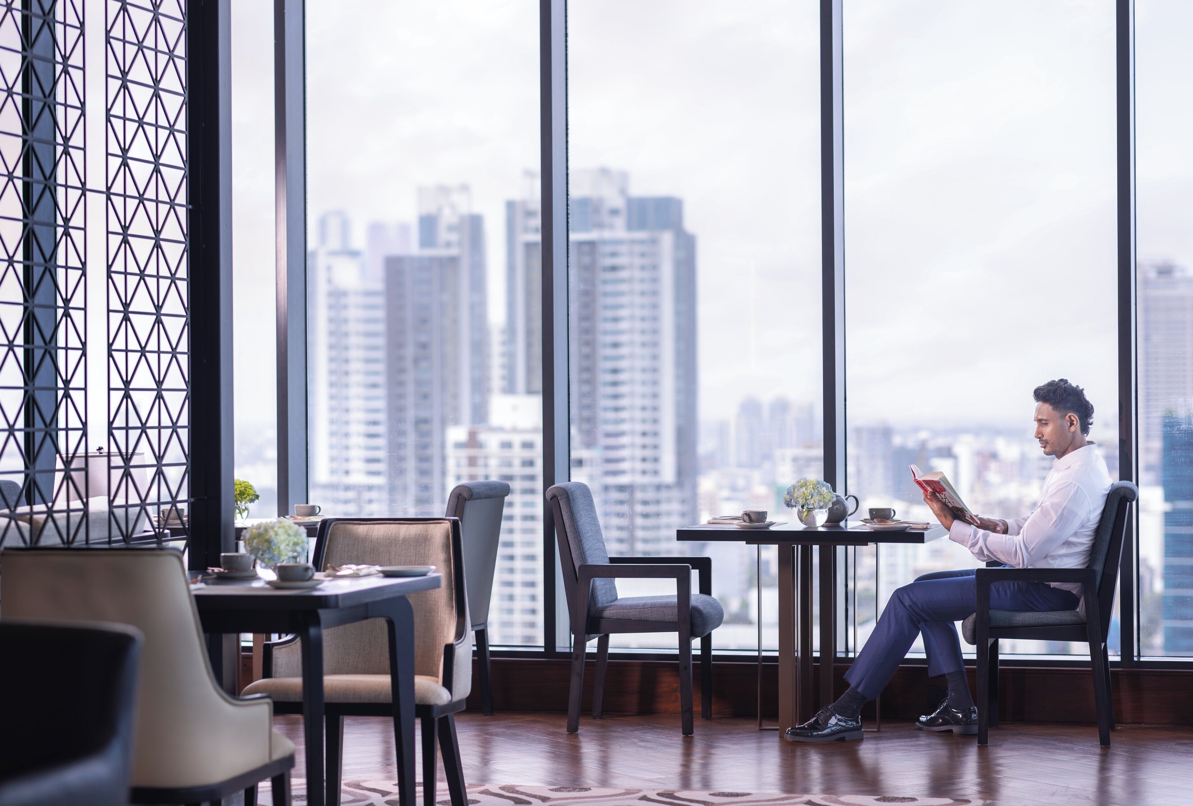 A man reading a book in a modern room with large windows, a city view, and a geometric patterned wall.