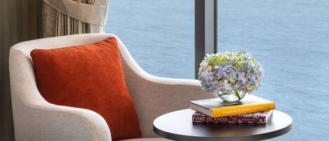 Una habitación con vista al mar, una silla con un cojín rojo, una mesita con libros y un florero con flores.