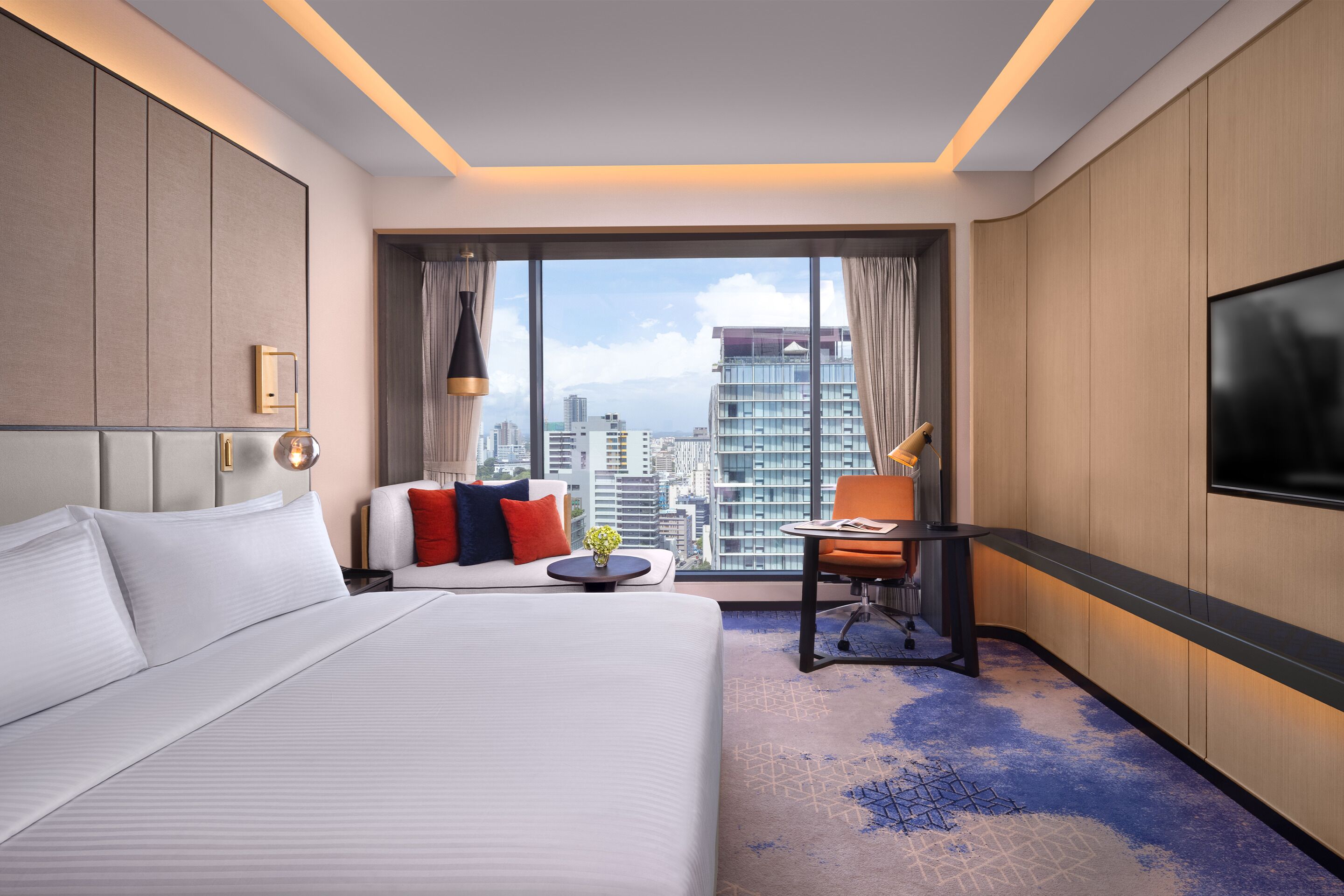 A hotel room with a large bed, a desk, a chair, and a TV. The room has a city view through large windows.