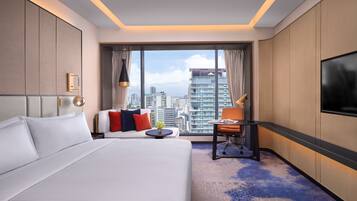 A hotel room with a large bed, a desk, a chair, and a TV. The room has a city view through large windows.