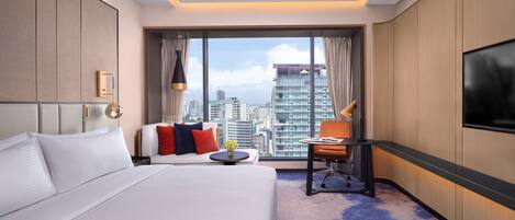 A hotel room with a large bed, a desk, a chair, and a TV. The room has a city view through large windows.