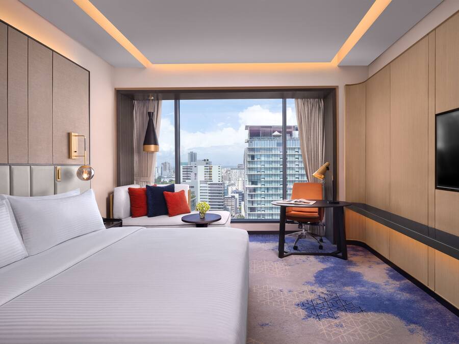 A hotel room with a large bed, a desk, a chair, and a TV. The room has a city view through large windows.