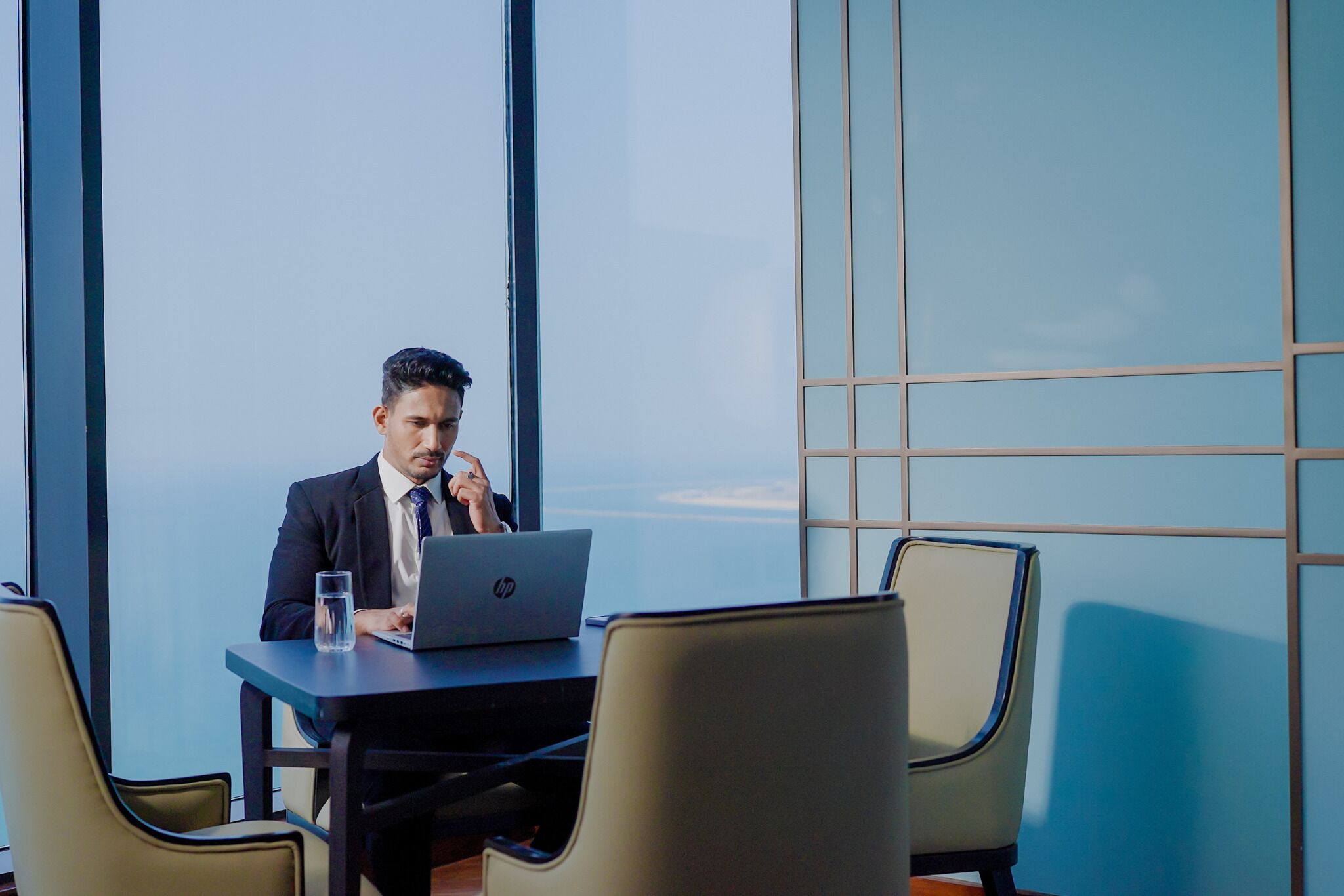 A man in a suit sitting at a table with a laptop, in a room with large windows and a view of the sky.