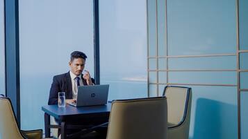 A man in a suit sitting at a table with a laptop, in a room with large windows and a view of the sky.