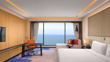 A hotel room with a bed, a desk, an orange chair, a sofa, a TV, and a view of the ocean.
