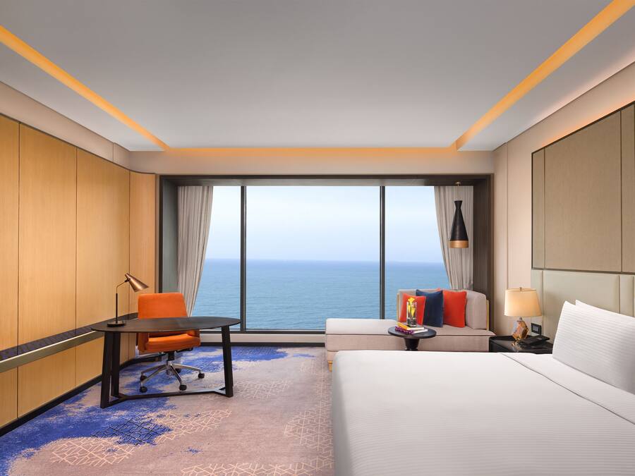A hotel room with a bed, a desk, an orange chair, a sofa, a TV, and a view of the ocean.