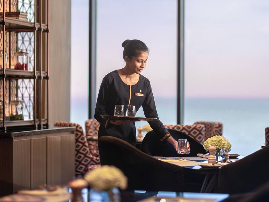 A restaurant with a view of the ocean, featuring a waiter serving a tray with glasses.