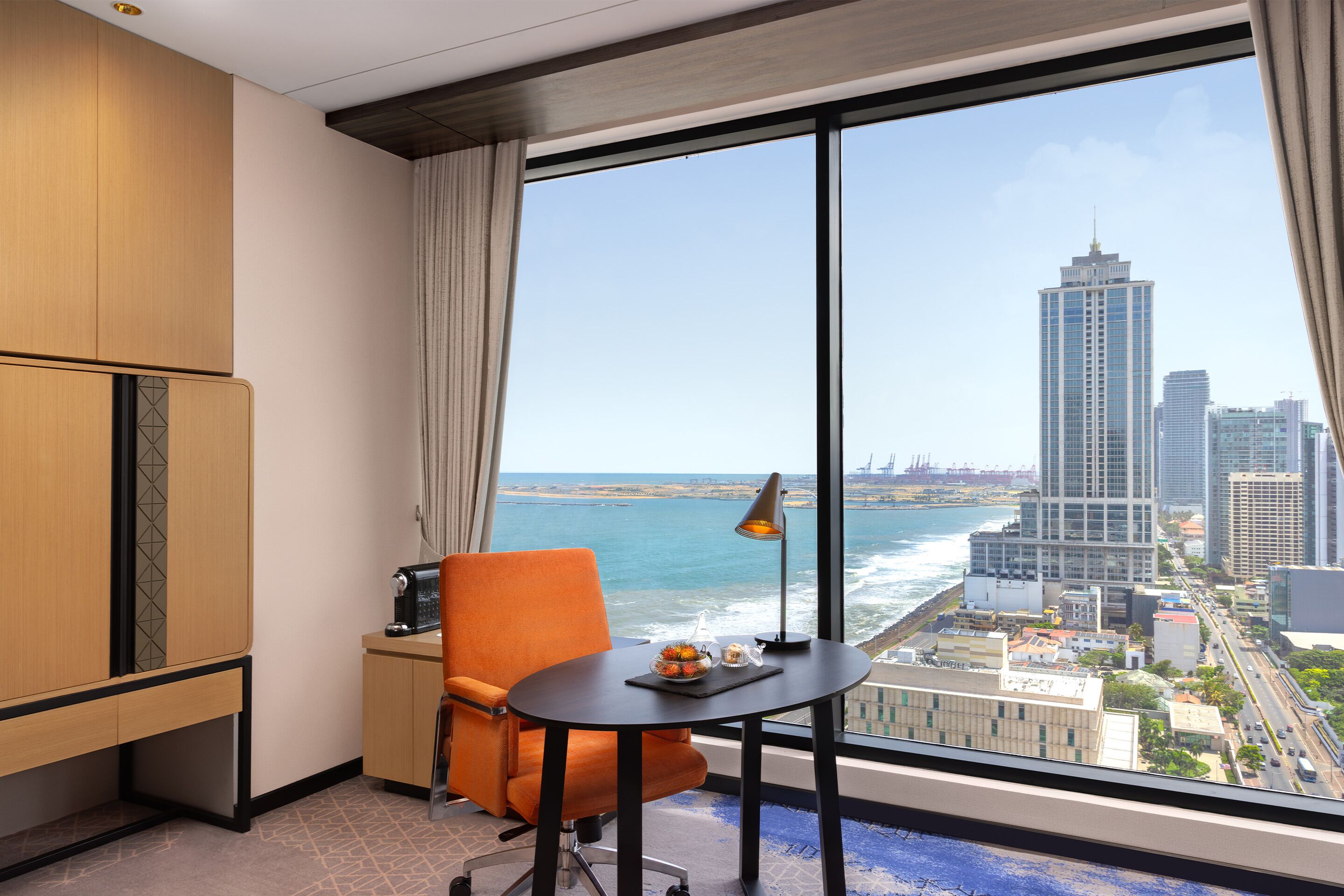 A hotel room with a large window, a view of the ocean and city skyline, a wooden wardrobe, an orange chair, and a small table with a lamp and a decorative item.