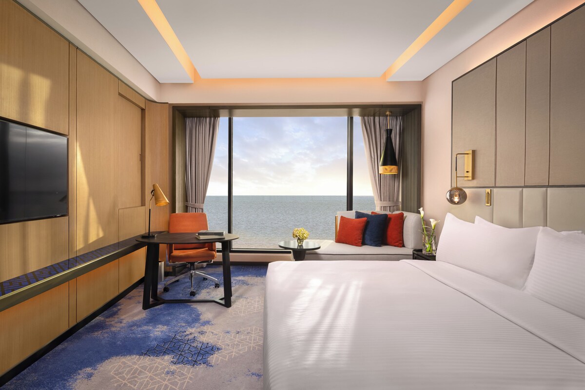 A hotel room with a large bed, a desk, a chair, a TV, and a view of the sea.