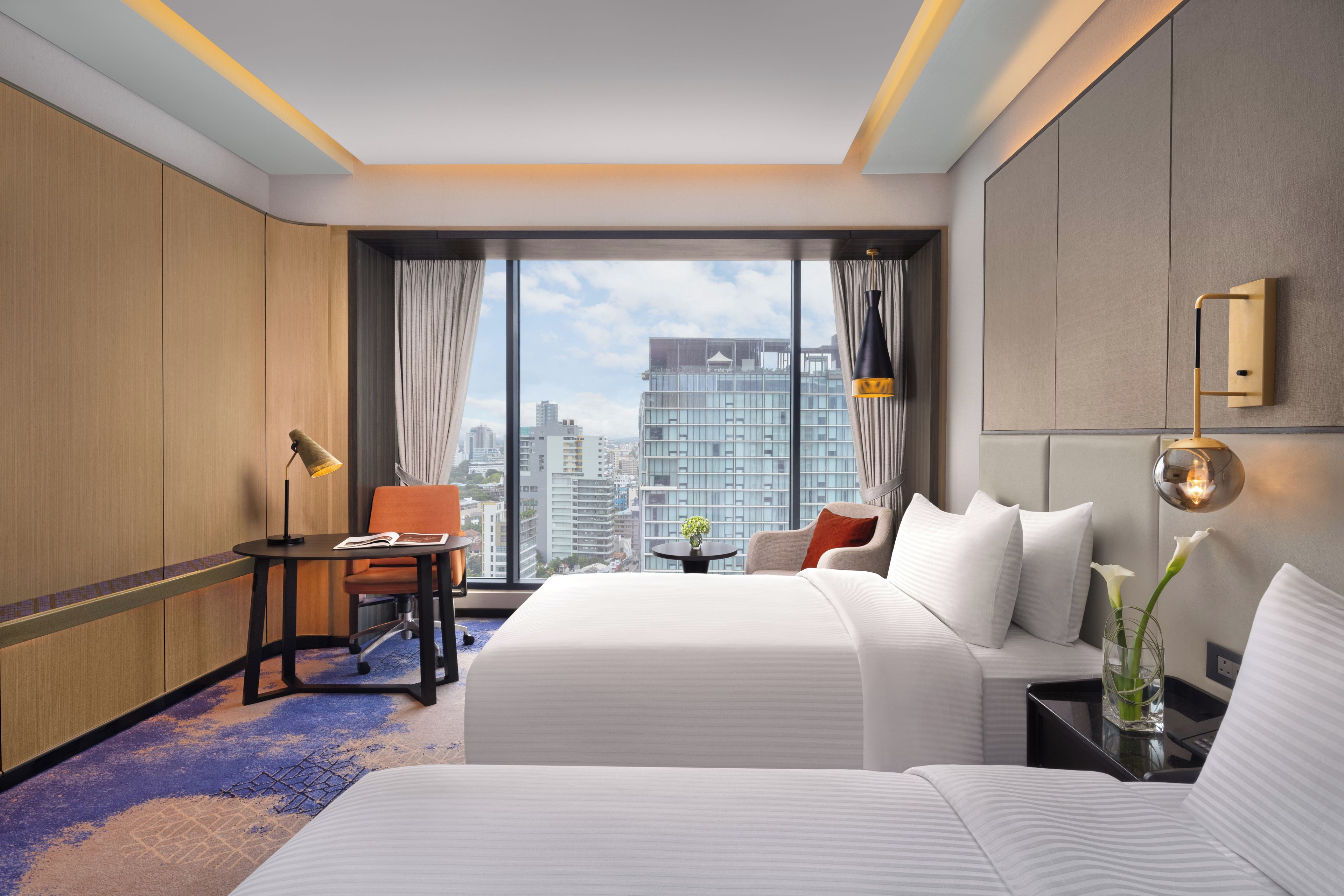 Een hotelkamer met een groot bed, een bureau, een stoel, een klein tafeltje met een vaas, en een uitzicht op de stad door grote ramen.