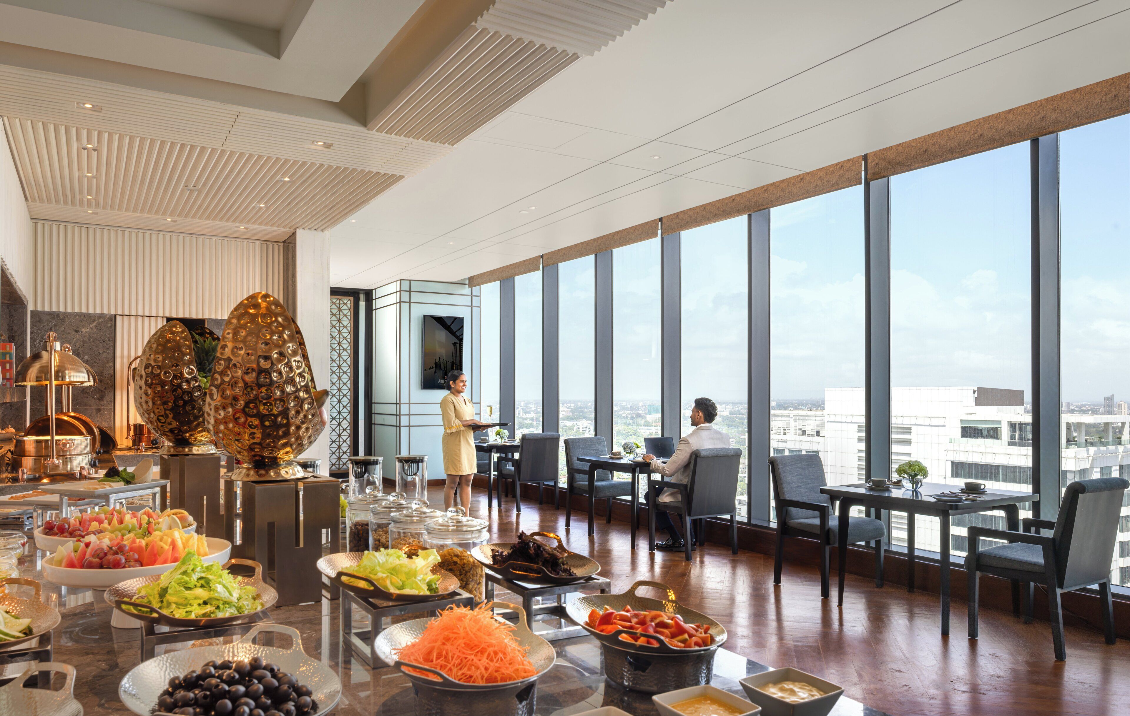 A high-rise hotel room with a panoramic city view, a buffet with various food items, and a modern interior design.