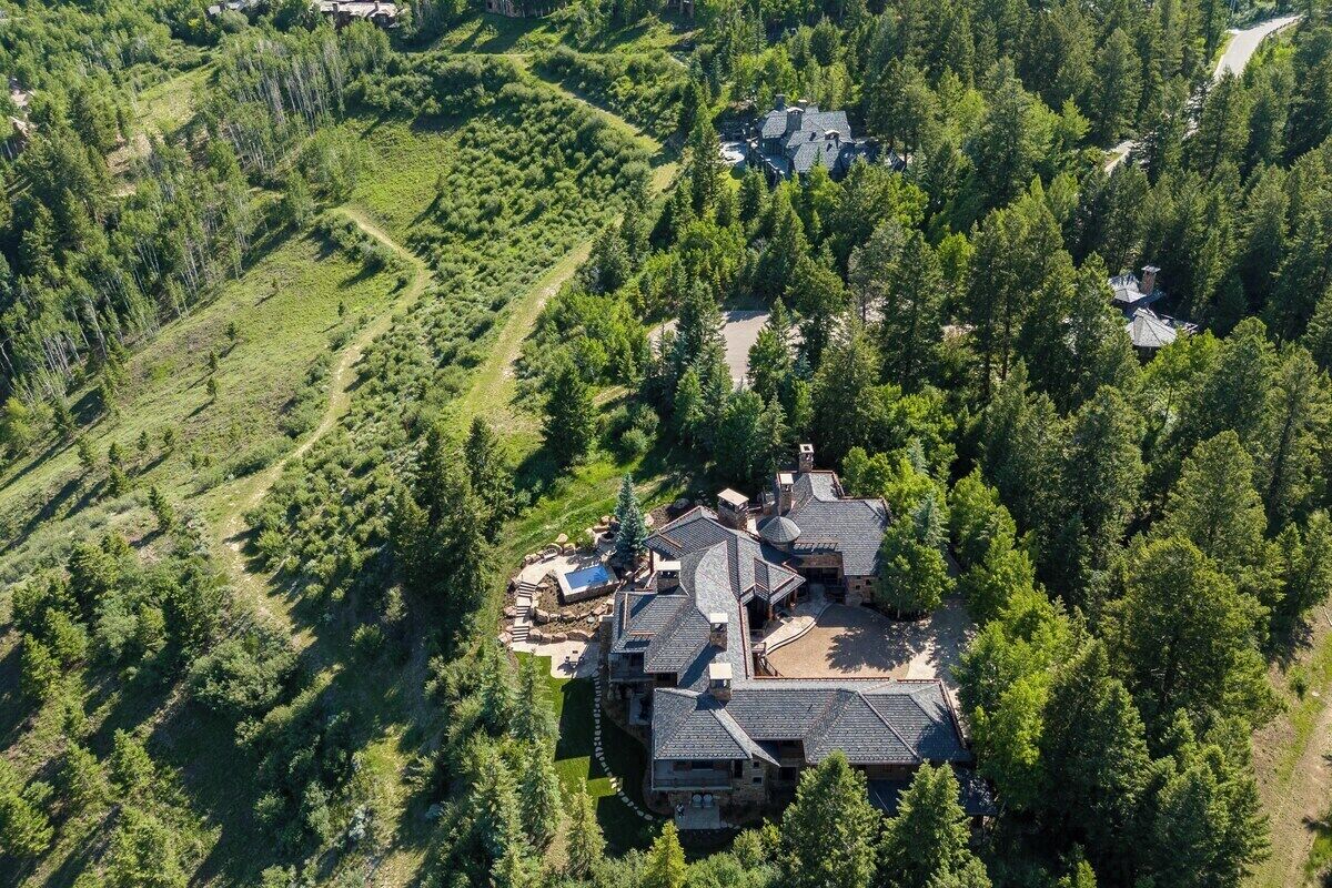 A large, multi-story house surrounded by dense forest, with a visible swimming pool and a well-maintained lawn.