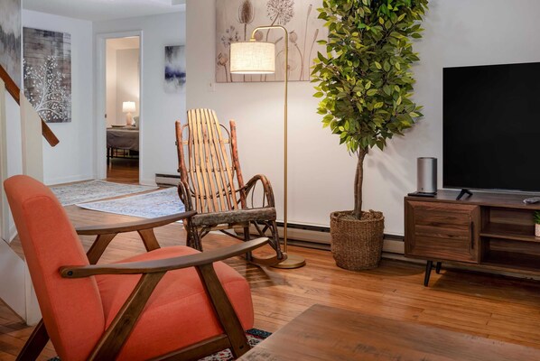 A living room with a wooden floor, a television, a plant, a rocking chair, and a lamp. - Cozy cabin w/hot tub & wraparound deck (Berkeley Springs)