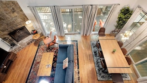 A spacious living area with a blue sofa, wooden coffee table, and a dining table with chairs. A stone wall and a large window with curtains are visible. - Cozy cabin w/hot tub & wraparound deck (Berkeley Springs)
