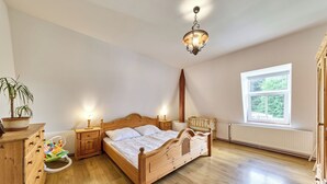 A bedroom with a wooden bed, bedside tables, a nightstand, a hanging light fixture, a window with a view, and a radiator.