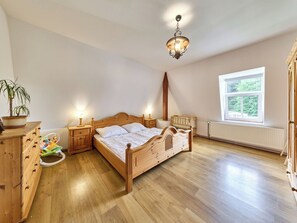 A bedroom with a wooden bed, bedside tables, a nightstand, a hanging light fixture, a window with a view, and a radiator.