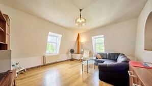 A spacious living room with a wooden floor, a black leather sofa, a glass coffee table, a white radiator, and two windows with a view of greenery.
