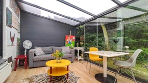 Un salon moderne avec un mur en verre, un canapé gris, une table jaune et un ventilateur blanc.