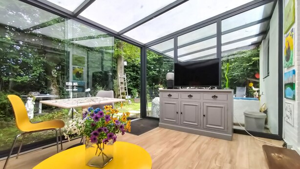 Un intérieur moderne avec une paroi vitrée, une table jaune ornée d’un vase de fleurs et un meuble gris sur lequel est posé un téléviseur à écran plat.