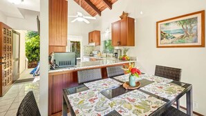 A modern kitchen with wooden cabinets, a central island, and a dining area with a table set for two. - Poipu Crater 24 – Stay in the Crater, A/C (Koloa)