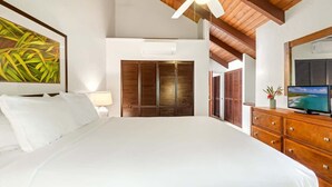 A bedroom with a large bed, wooden ceiling beams, a ceiling fan, a TV, and a wardrobe. - Poipu Crater 23 – Vacation in the Crater, AC in Bedrooms (Koloa)