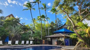 A pool area with lounge chairs, a blue umbrella, and a building with a tiled roof. - Waikomo Stream Villas 103 – Ground Floor, Garden View Retreat (Koloa)
