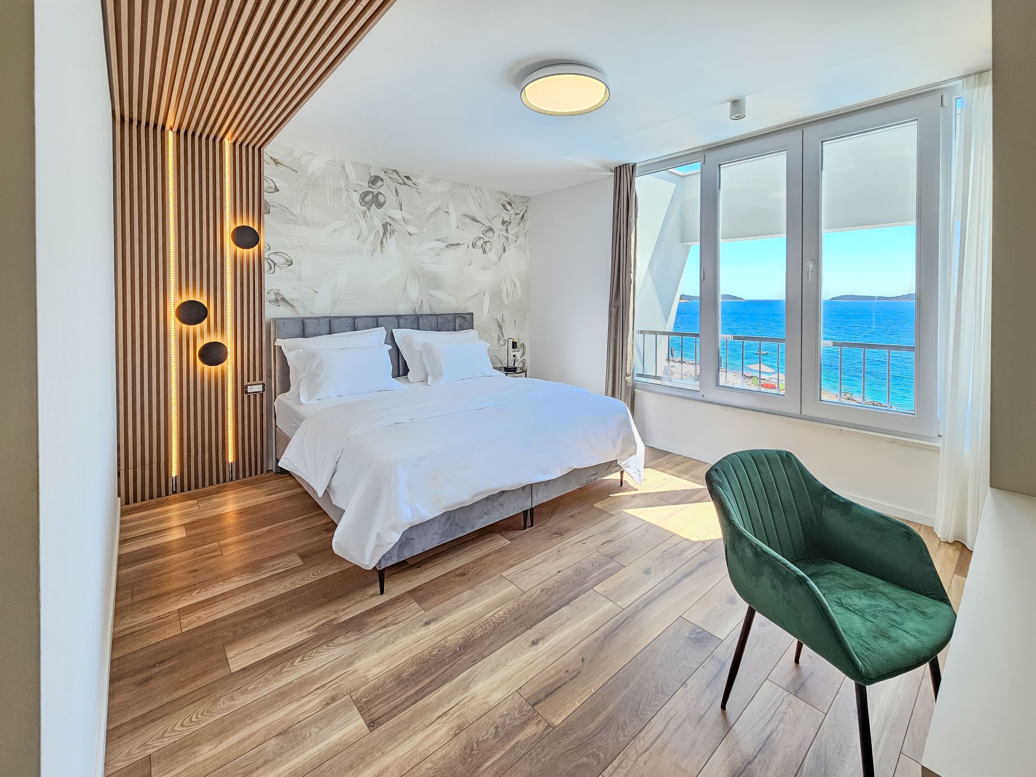 A modern bedroom with a large bed, a green chair, and a view of the sea through large windows.