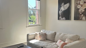 Una sala de estar con un sofá seccional color beige, una mesa de centro de madera, un ventilador de techo, una ventana con vistas a la vegetación y dos obras de arte enmarcadas en la pared.