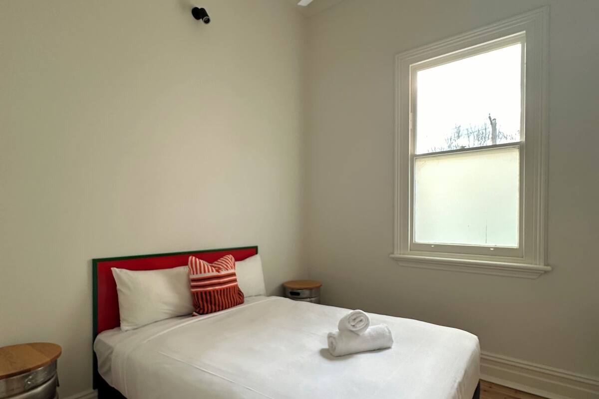 Cama bem arrumada com lençóis brancos, cabeceira vermelha e almofadas listradas. Piso de madeira, ventilador de teto e janela com vista para o céu.