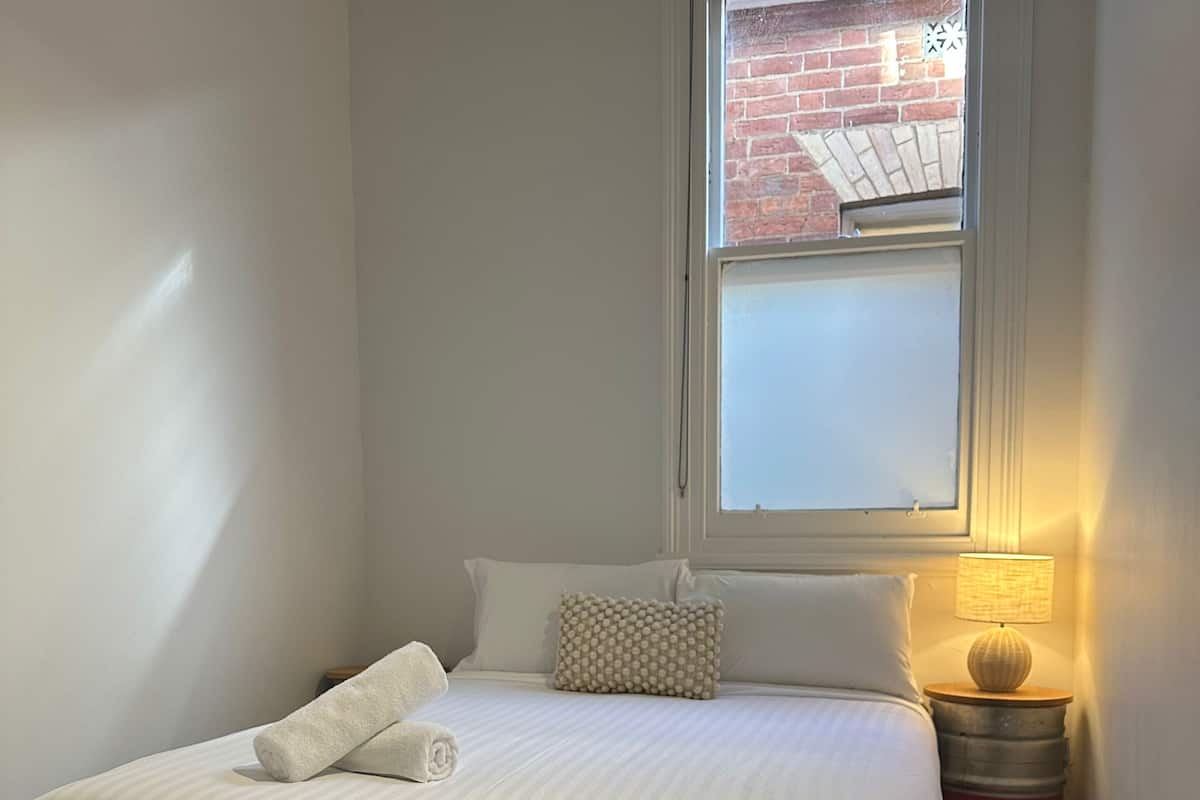 Uma cama bem arrumada com lençóis brancos, uma luminária de cabeceira e uma janela com vista para um muro de tijolos.