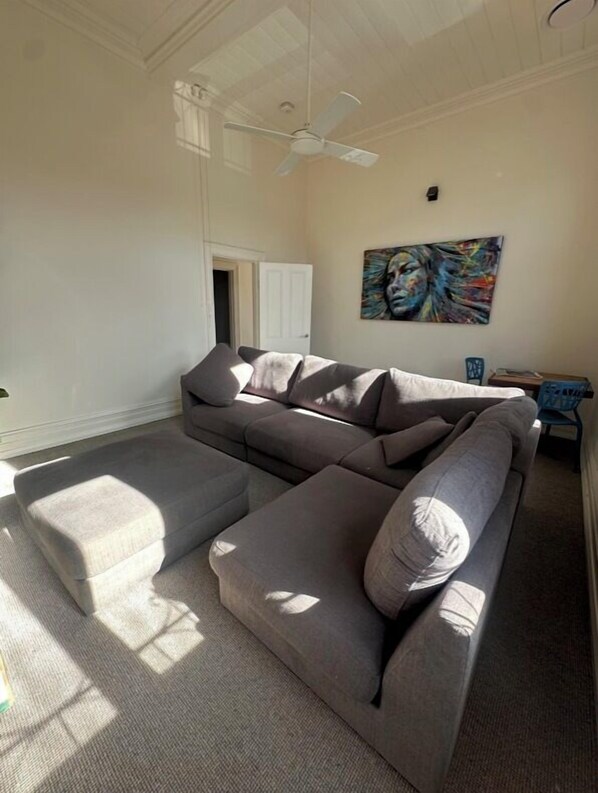 A living room with a sectional sofa, a ceiling fan, and a painting on the wall. - ARK Apartments (Echuca)