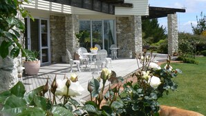 Une maison moderne avec une façade en pierre, un patio doté de mobilier d’extérieur et un jardin agrémenté de rosiers et d’autres plantes.