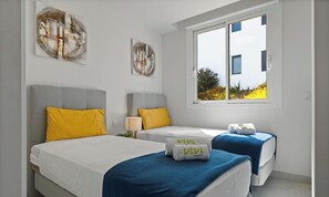 A hotel room with two beds, a window with a view of greenery, and artwork on the walls.