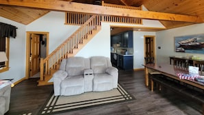 Ein großzügiger Wohnbereich mit hohen Decken, einer Holztreppe, einem grauen Sofa und einer Küche im Hintergrund.