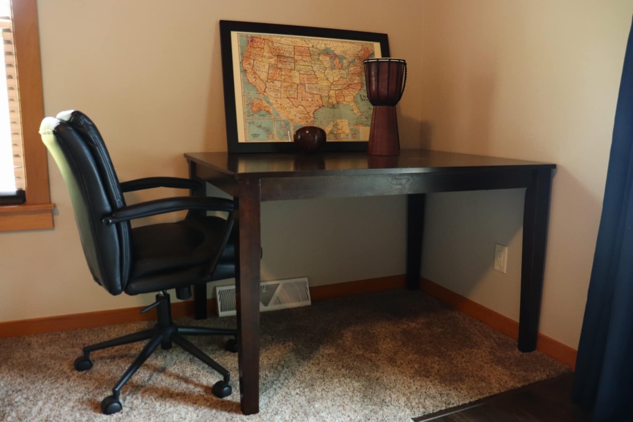 A desk with a framed map, a black office chair, and a decorative vase.