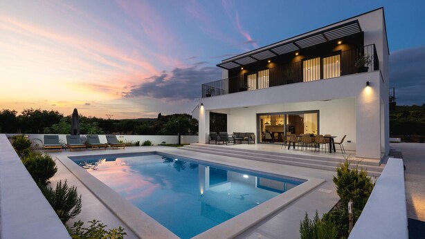 Una casa moderna de dos pisos con piscina, mobiliario exterior y vistas a la puesta de sol.